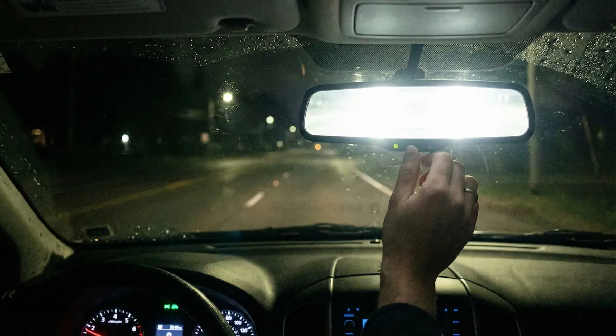 Visão do motorista dentro do carro à noite com chuva, mostrando o espelho retrovisor refletindo um farol alto cegante e uma mão acionando a alavanca do modo noturno.